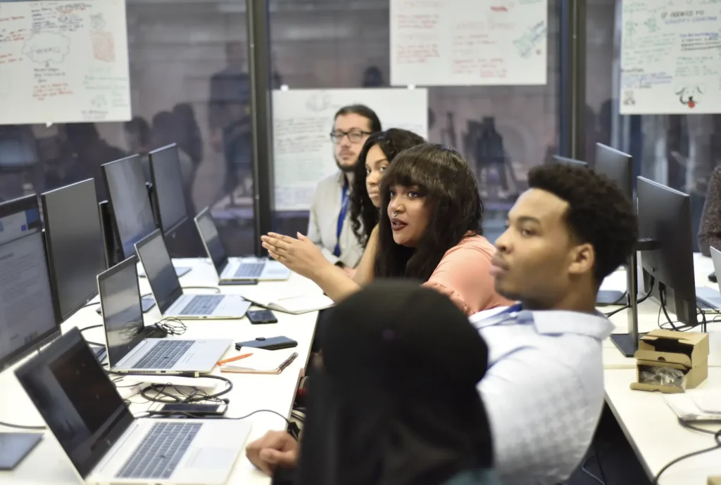 Individuals sat in a row at a white long desk with laptops sitting on the table.
